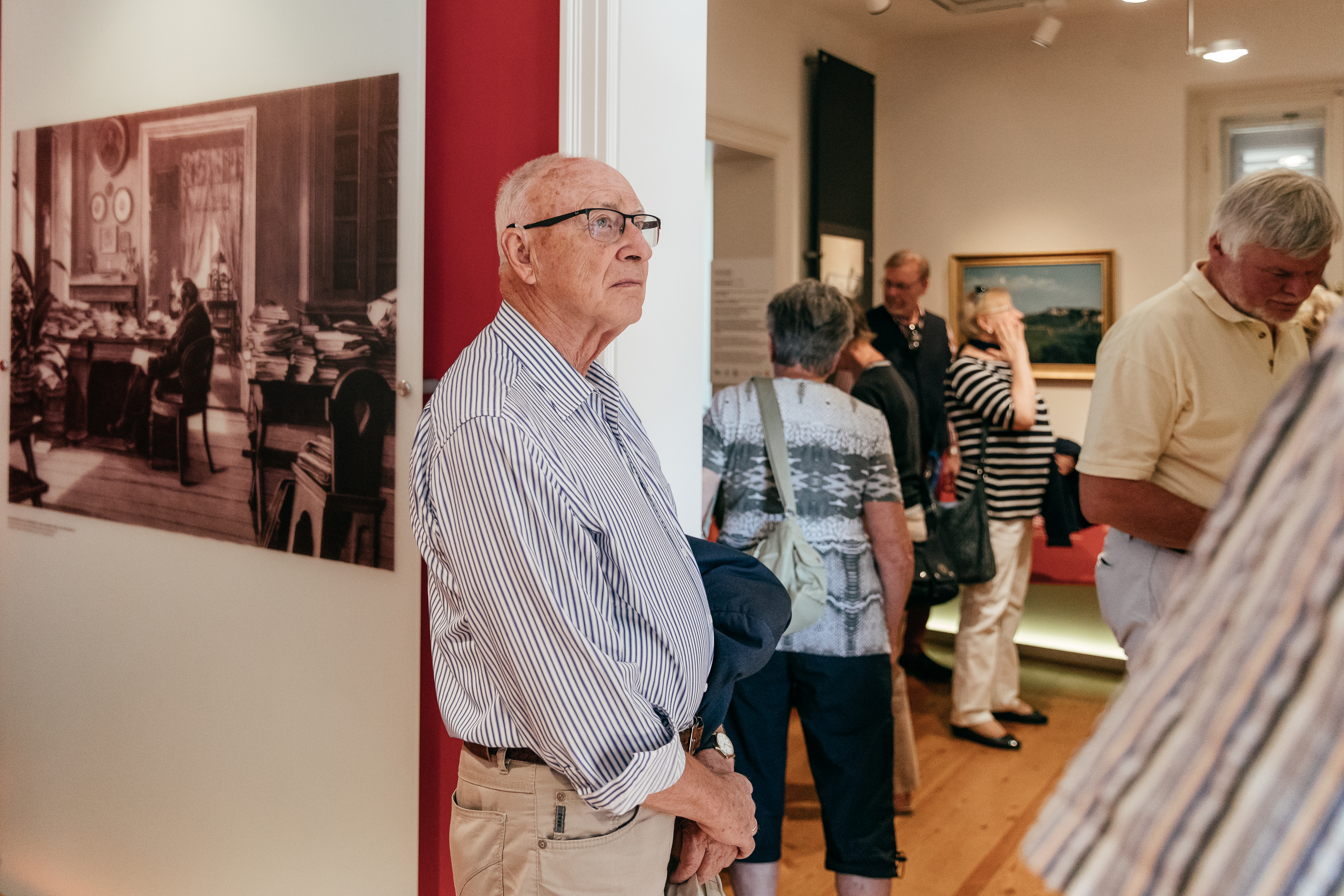 KI generiert: Das Bild zeigt mehrere Personen, die sich in einem Ausstellungsraum befinden. Ein älterer Mann steht im Vordergrund, wobei im Hintergrund weitere Besucher sowie Gemälde und Fotografien an den Wänden zu sehen sind.