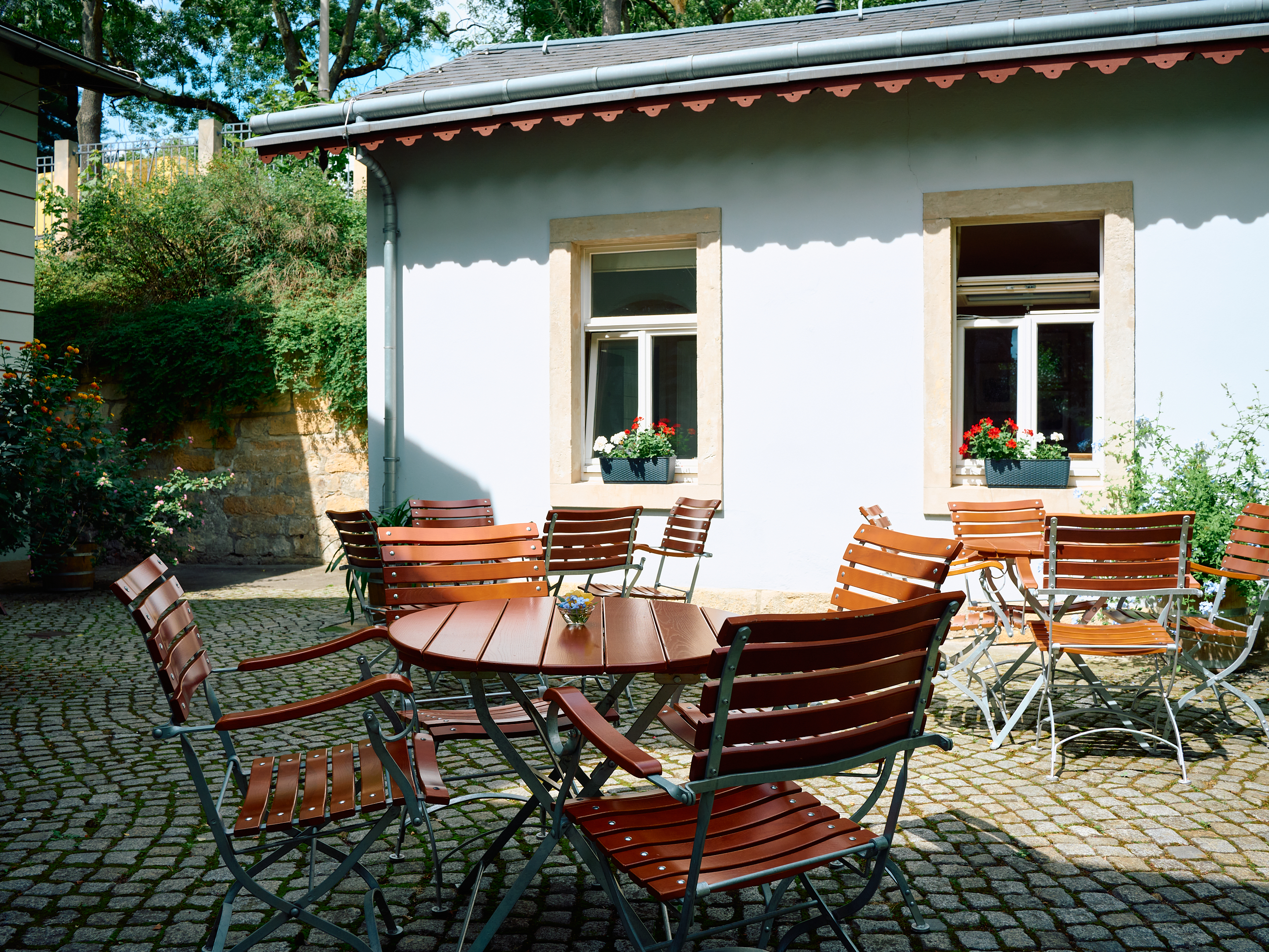 KI generiert: Das Bild zeigt einen Außenbereich eines gemütlichen Cafés oder Restaurants mit mehreren leeren Holztischen und -stühlen auf einem gepflasterten Untergrund. Im Hintergrund sieht man eine Hauswand mit zwei Fenstern, die mit Blumenkästen geschmückt sind.