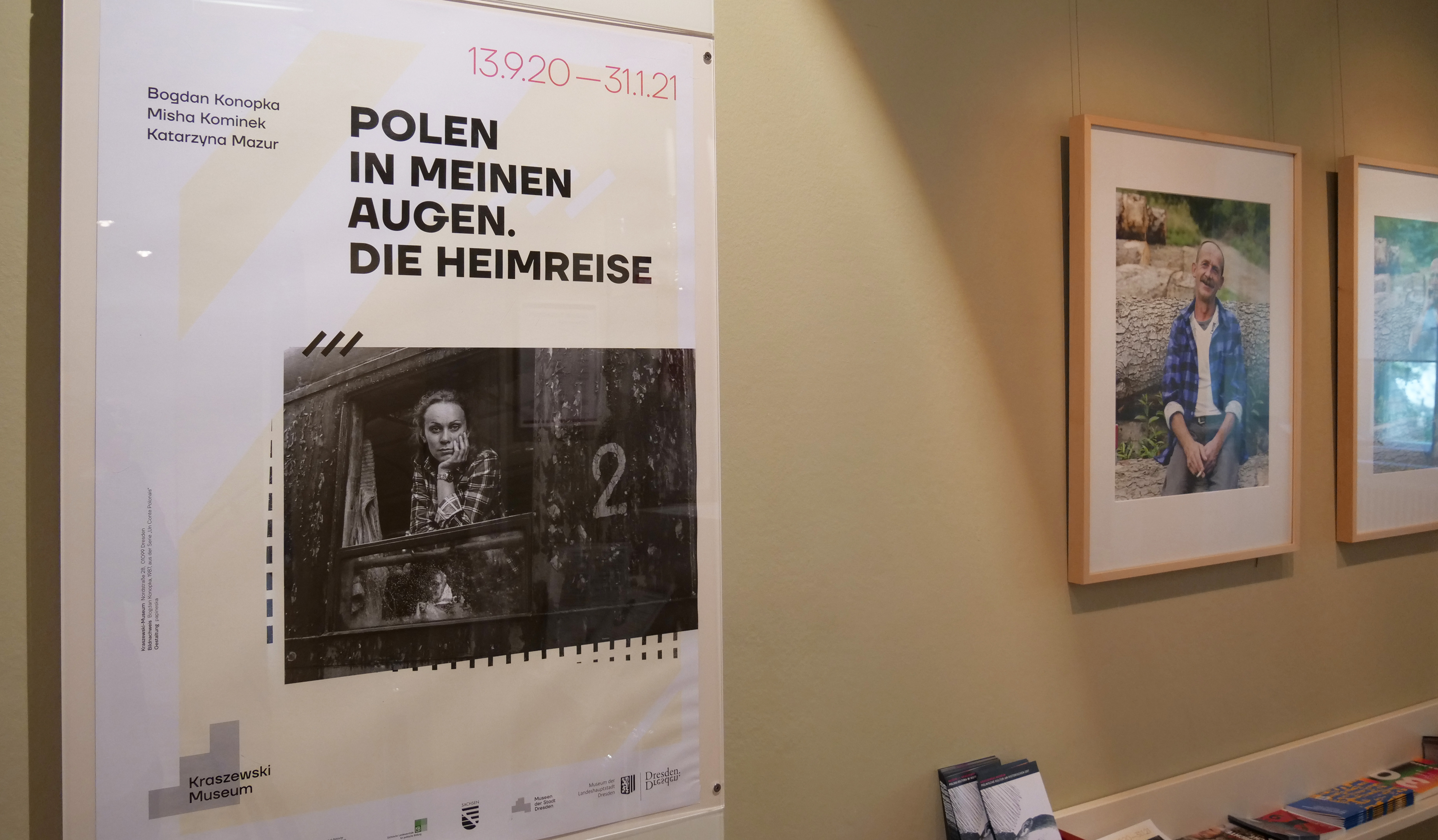 KI generiert: Das Bild zeigt ein Plakat für eine Ausstellung mit dem Titel "Polen in meinen Augen. Die Heimreise." im Kraszewski Museum sowie zwei gerahmte Fotos an der Wand daneben.