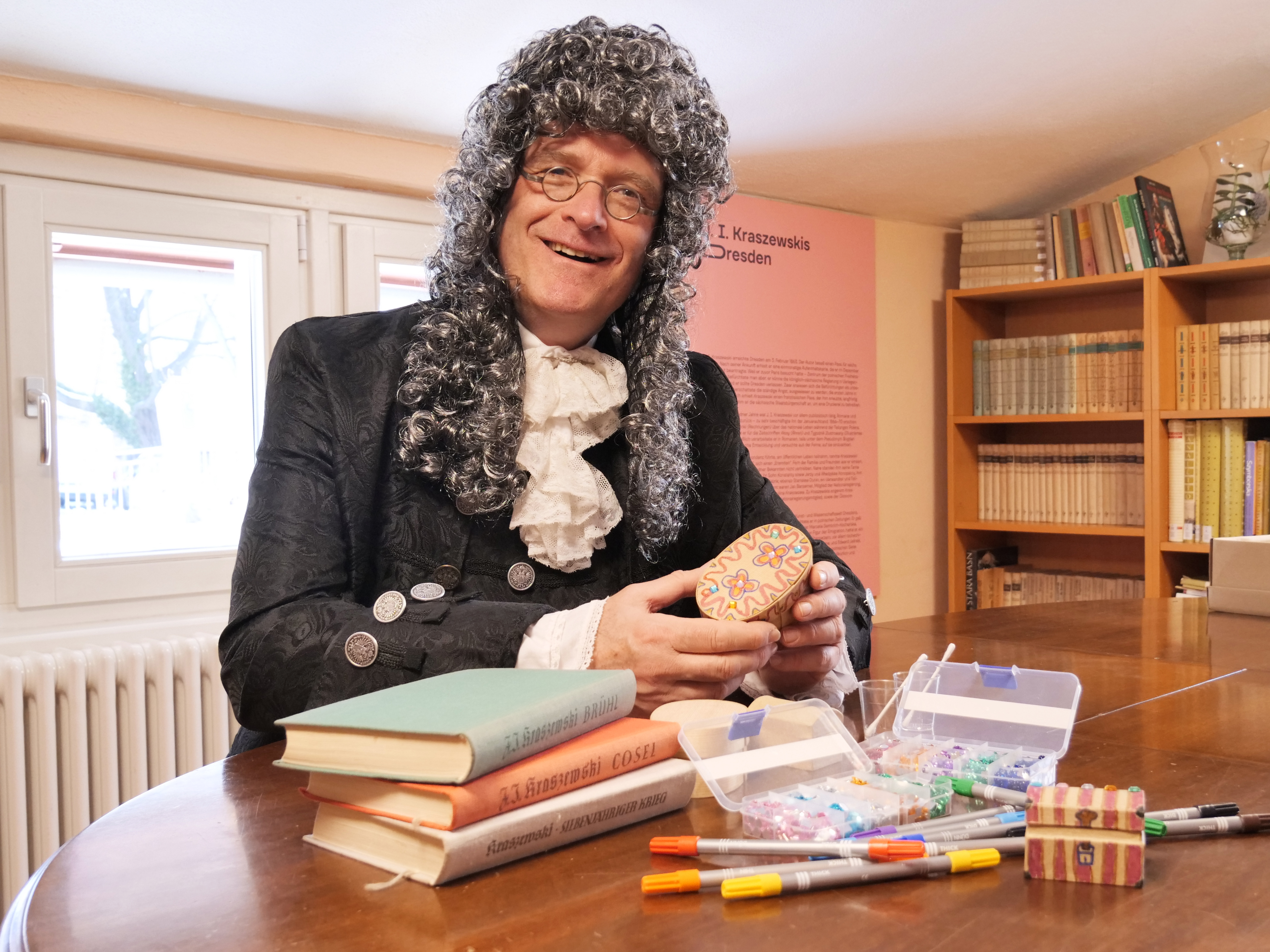 KI generiert: Ein Mann in historischer Kleidung sitzt an einem Tisch, hält ein bunt bemaltes Ei in der Hand und ist von Büchern und Malutensilien umgeben. Der Raum wirkt wie eine Bibliothek oder ein Arbeitszimmer.