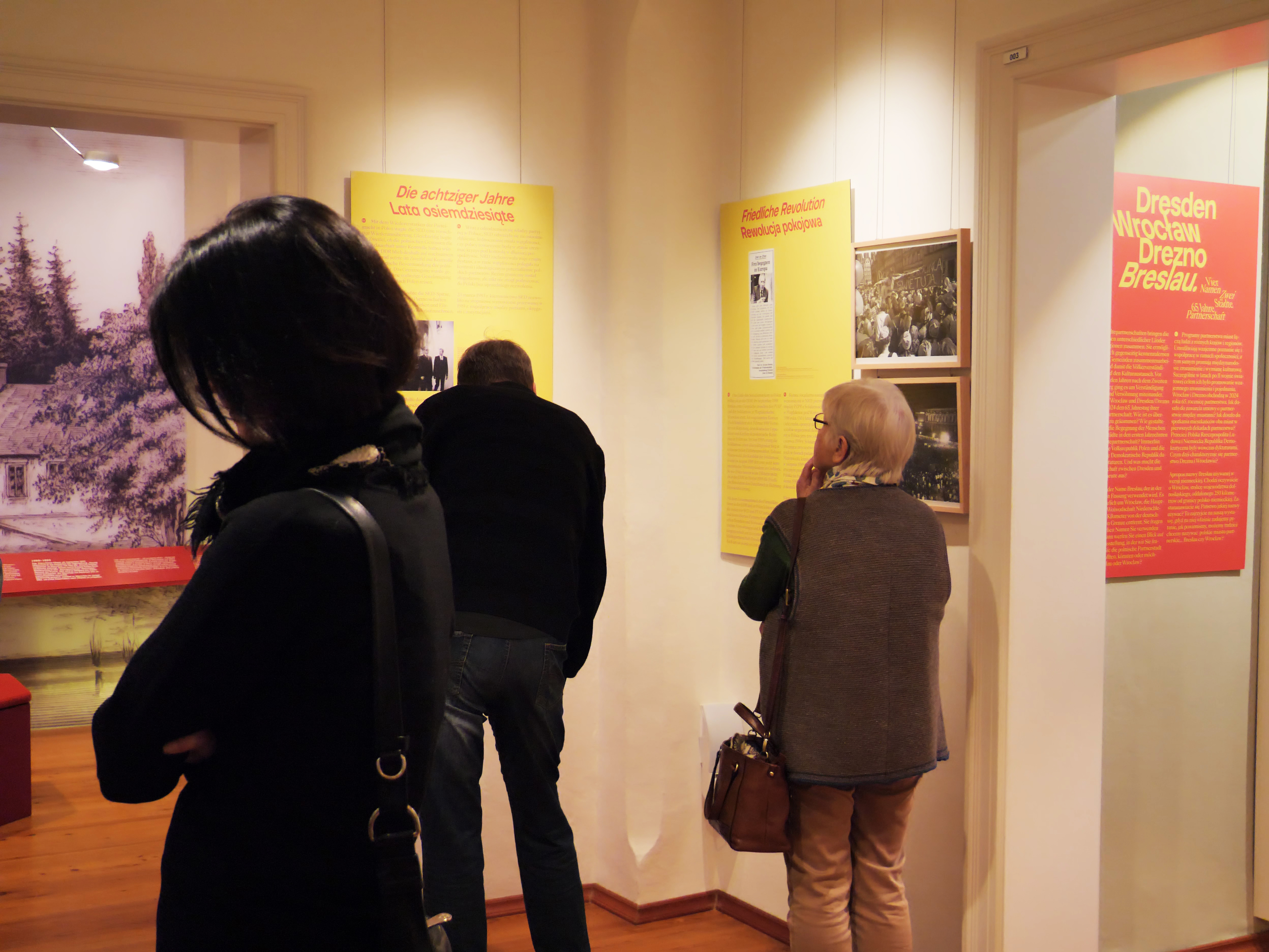 KI generiert: Das Bild zeigt Menschen in einem Museum oder einer Ausstellung, die sich informative Tafeln und Bilder an den Wänden ansehen. Die Ausstellung scheint sich mit historischen Themen zu beschäftigen, möglicherweise mit Bezug zu Dresden.