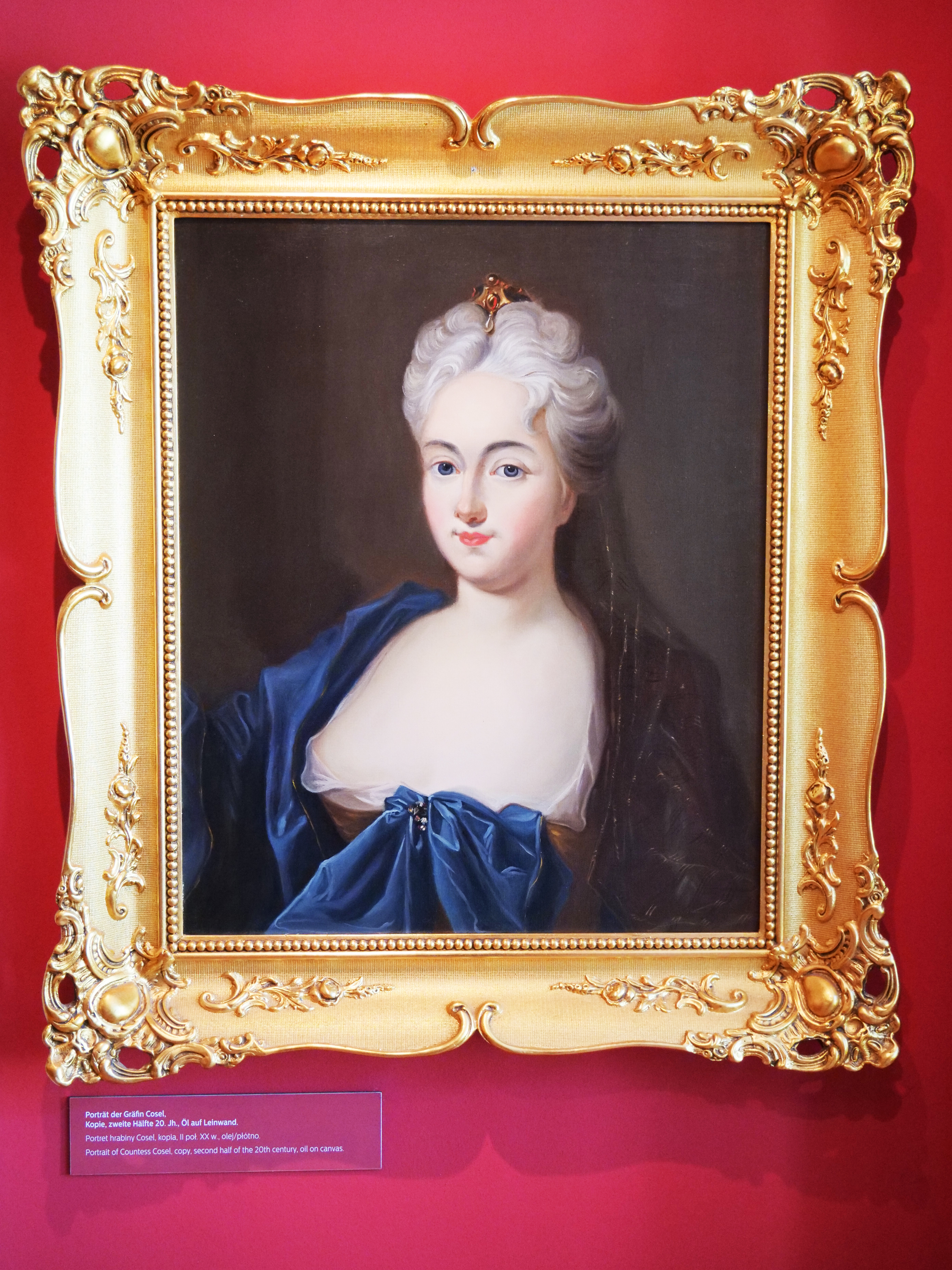 KI generiert: Das Bild zeigt ein klassisches Gemälde einer Frau in einem prunkvollen, goldenen Rahmen vor einem roten Hintergrund. Die Frau trägt ein blaues Kleid und hat eine elegante Frisur.