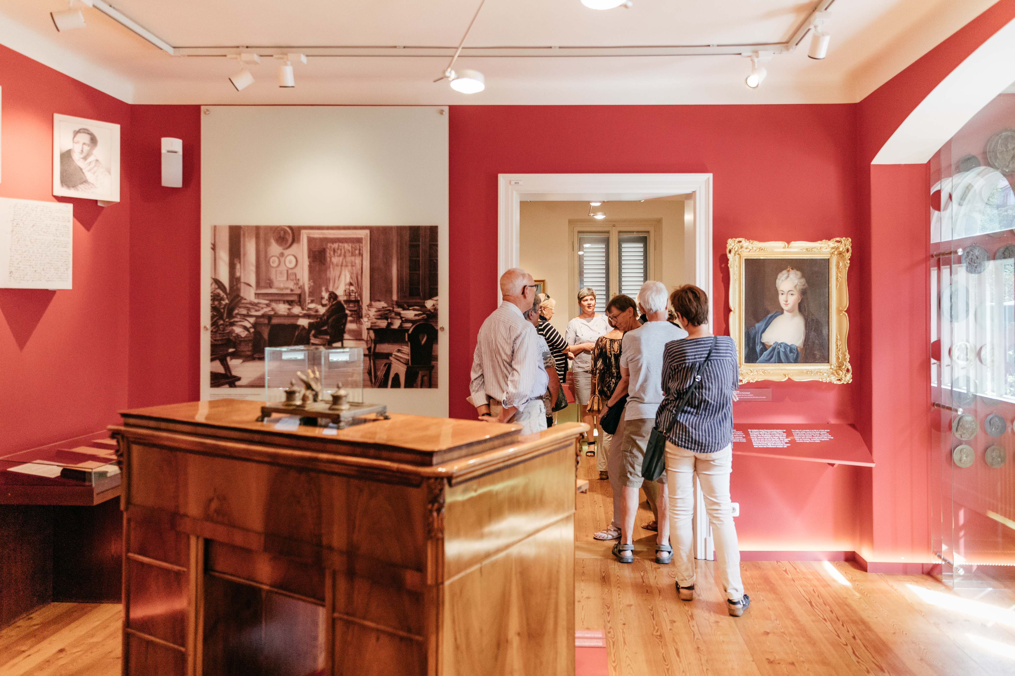 KI generiert: Das Bild zeigt eine Gruppe von Menschen, die sich ein Museum oder eine Ausstellung mit historischen Fotografien und Gemälden ansehen. Die Wände des Raumes sind rot gestrichen und es sind mehrere Kunstwerke und Exponate zu sehen.