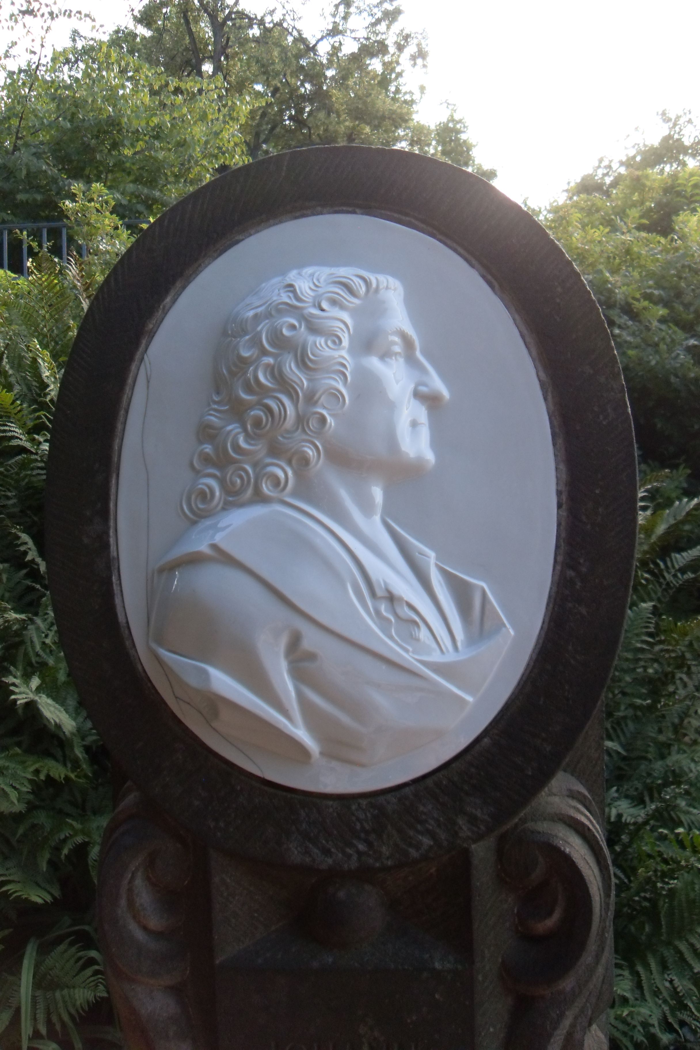 AI generated: The image shows a relief portrait of a person in profile view, embedded in a decorative, oval frame relief on a plinth. There are plants and trees in the background.
