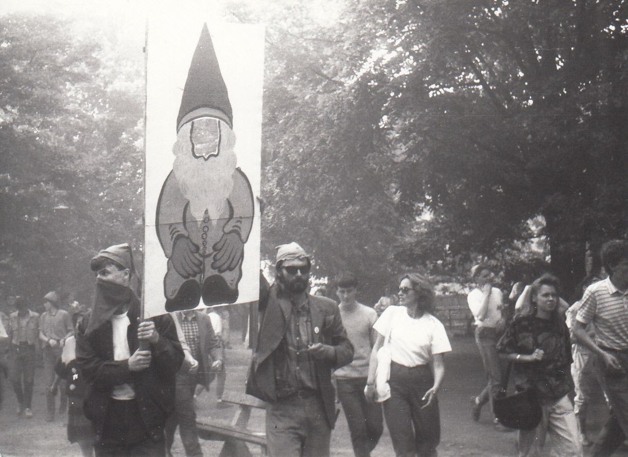 KI generiert: Das Bild zeigt eine Gruppe von Menschen bei einer Demonstration, wobei zwei Personen ein großes Plakat mit einem Bild eines Gartenwichtels tragen. Die Szenerie ist in Schwarz-Weiß gehalten und vermittelt eine lebhafte Proteststimmung.