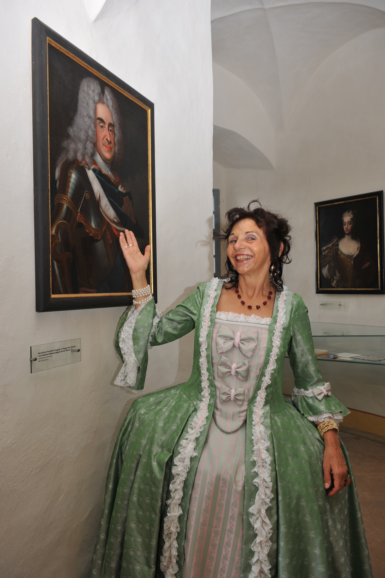 KI generiert: Eine Frau in einem grünen, historischen Kostüm präsentiert lächelnd ein Porträt eines Mannes an der Wand in einem Ausstellungsraum.